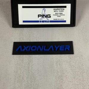 May include: Black rectangular label with the text "AXIONLAYER" in blue. Above it is a black rectangular case with a white label that reads "PING ZZ LITE" and "KARSTEN MFG. CORP." in black text. Made in U.S.A.