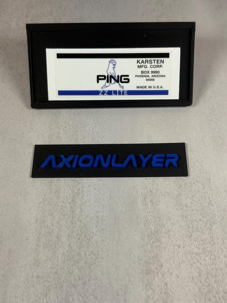 May include: Black rectangular label with the text "AXIONLAYER" in blue. Above it is a black rectangular case with a white label that reads "PING ZZ LITE" and "KARSTEN MFG. CORP." in black text. Made in U.S.A.