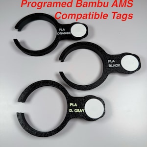 May include: Three black circular tags with open gaps, labeled Programed Bambu AMS Compatible Tags. Each tag features a white circular insert and text reading PLA ORANGE, PLA BLACK, or PLA D. GRAY, designed for 3D printer filament identification.