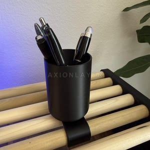 May include: A matte black cylindrical pen holder with a curved clip attachment, secured onto a light-coloured wooden slat rack. The holder contains several black pens with silver accents. The text AXIONLAYER is visible on the holder surface.