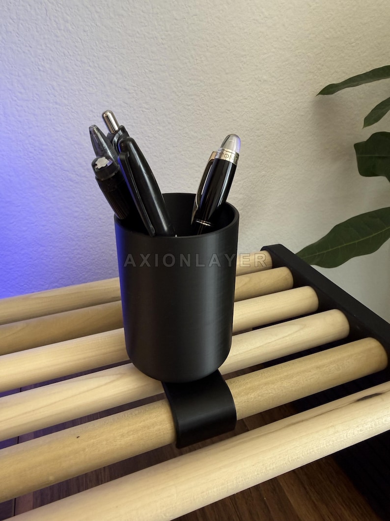 May include: A matte black cylindrical pen holder with a curved clip attachment, secured onto a light-coloured wooden slat rack. The holder contains several black pens with silver accents. The text AXIONLAYER is visible on the holder surface.