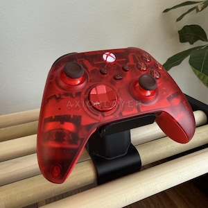 May include: A translucent red wireless video game controller featuring the text AXIONLAYER printed on the front. The controller has two dark brown analogue sticks, a red directional pad, a glowing white Xbox button, and standard action buttons.