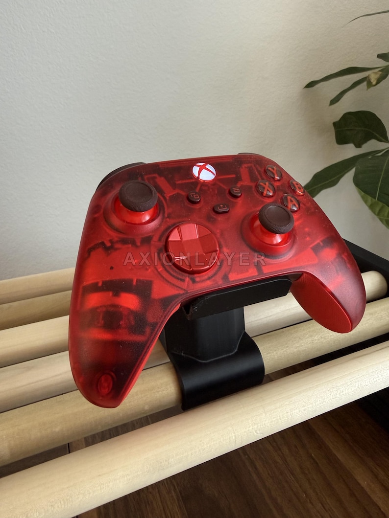 May include: A translucent red wireless video game controller featuring the text AXIONLAYER printed on the front. The controller has two dark brown analogue sticks, a red directional pad, a glowing white Xbox button, and standard action buttons.