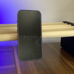 May include: A black smartphone with a dark screen featuring the text AXIONLAYER in the centre. The device is positioned vertically, resting against a light-coloured wooden slat structure. The phone has a slim frame and a rounded rectangular shape.