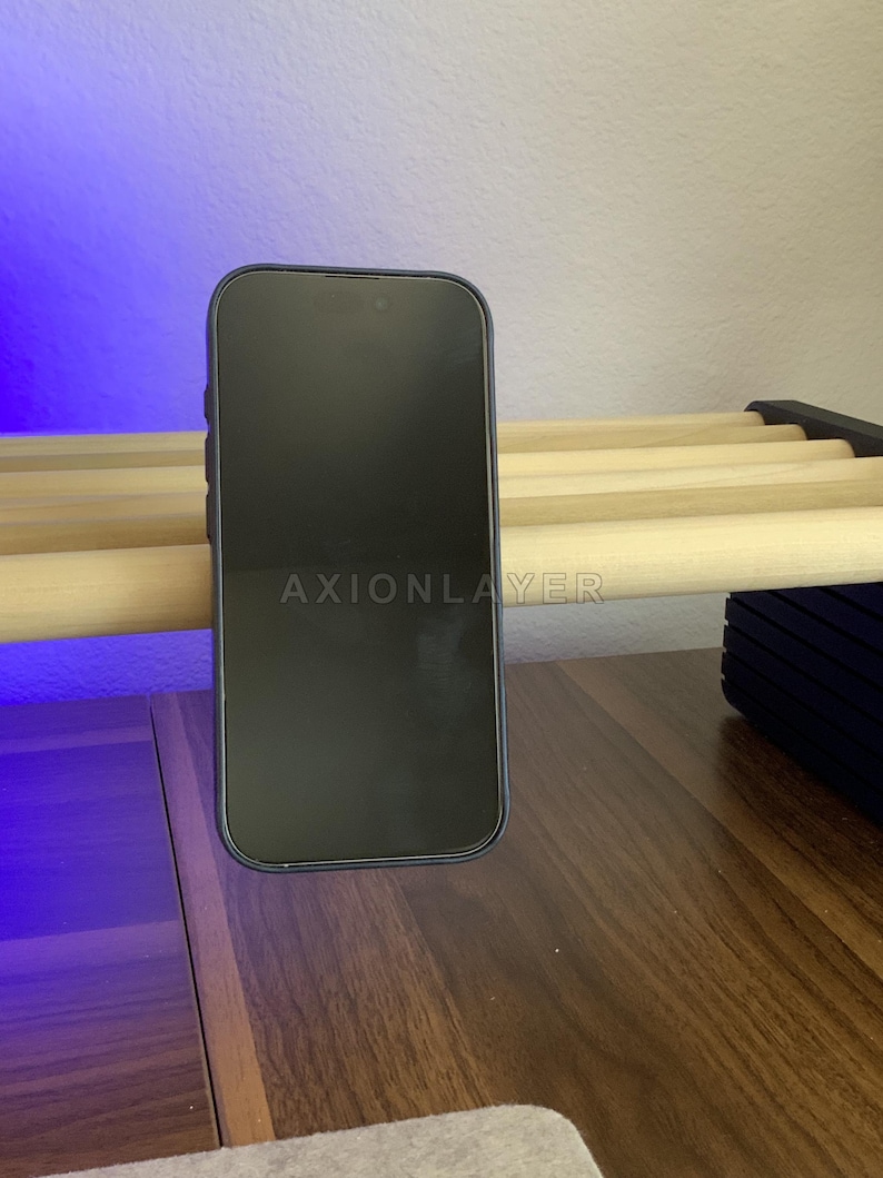 May include: A black smartphone with a dark screen featuring the text AXIONLAYER in the centre. The device is positioned vertically, resting against a light-coloured wooden slat structure. The phone has a slim frame and a rounded rectangular shape.
