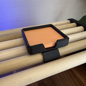 May include: A black square plastic holder with a clip attachment, featuring the text AXIONLAYER printed on the front. The holder contains a stack of orange square sticky notes and is clipped onto a light-coloured wooden dowel rack.