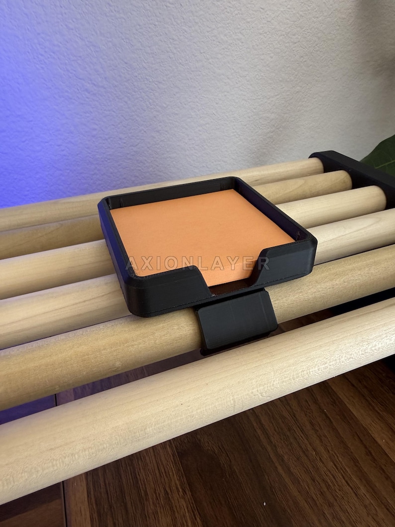 May include: A black square plastic holder with a clip attachment, featuring the text AXIONLAYER printed on the front. The holder contains a stack of orange square sticky notes and is clipped onto a light-coloured wooden dowel rack.