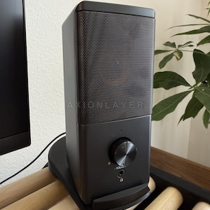 May include: A black rectangular speaker with a metal mesh grille on the upper half and the text AXIONLAYER printed on the front. The lower section features a circular volume control knob, a small indicator light, and a headphone jack port.