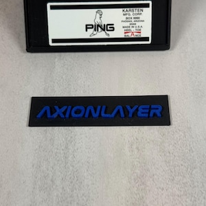 May include: Black rectangular item with the word "AXIONLAYER" in blue lettering. Above it is a black rectangular box with a white label that reads "PING" and other text including "MADE IN U.S.A."