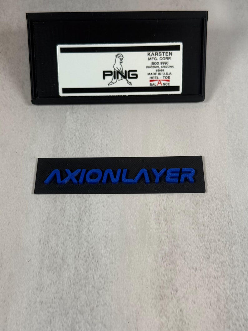 May include: Black rectangular item with the word "AXIONLAYER" in blue lettering. Above it is a black rectangular box with a white label that reads "PING" and other text including "MADE IN U.S.A."