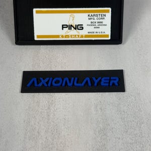 May include: Black rectangular label with the blue text "AXIONLAYER" and a black rectangular box with the text "PING" and "KT-SHAFT". The box also includes the text "KARSTEN MFG. CORP." and "MADE IN U.S.A."