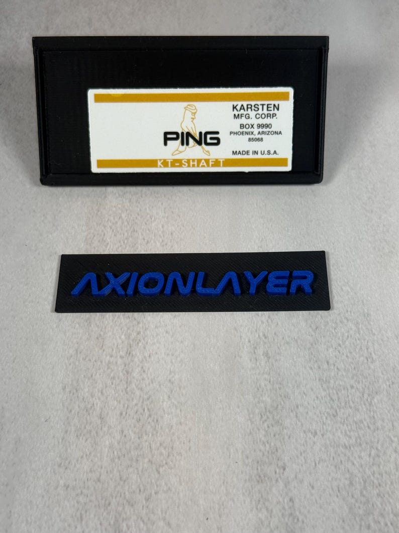 May include: Black rectangular label with the blue text "AXIONLAYER" and a black rectangular box with the text "PING" and "KT-SHAFT". The box also includes the text "KARSTEN MFG. CORP." and "MADE IN U.S.A."