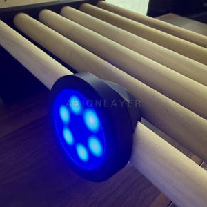 May include: A black circular device with a bright blue LED light ring featuring seven glowing dots and the text AXIONLAYER printed in white. The device is attached to a series of parallel, light-coloured wooden cylindrical rods arranged in a row.