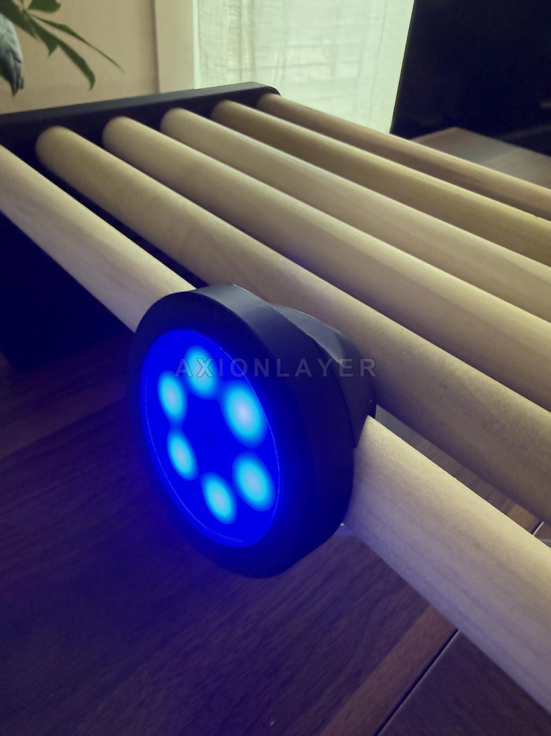 May include: A black circular device with a bright blue LED light ring featuring seven glowing dots and the text AXIONLAYER printed in white. The device is attached to a series of parallel, light-coloured wooden cylindrical rods arranged in a row.