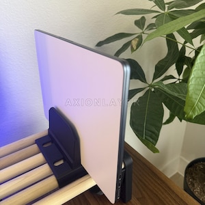 May include: A black plastic stand holding a thin, light-coloured tablet upright. The stand features a series of parallel light-coloured wooden dowels extending horizontally. The word AXIONLAYER is printed in white on the surface of the tablet.
