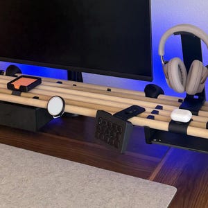 May include: A modular desk organiser featuring a black frame with horizontal light wood dowels. It includes a black storage drawer, a headphone stand, a tablet holder, a magnetic charger, and a numeric keypad mount, supporting a computer monitor.