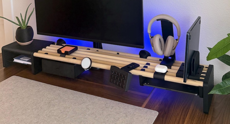May include: A modular desk organiser featuring a black frame with horizontal light wood dowels. It includes a black storage drawer, a headphone stand, a tablet holder, a magnetic charger, and a numeric keypad mount, supporting a computer monitor.
