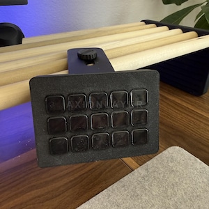 May include: A black macro keypad featuring a grid of fifteen square buttons with the text AXIONLAYER printed across the centre. The device includes a mounting bracket with a circular adjustment knob, designed to attach to a desk or shelf edge.