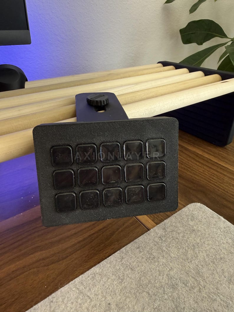 May include: A black macro keypad featuring a grid of fifteen square buttons with the text AXIONLAYER printed across the centre. The device includes a mounting bracket with a circular adjustment knob, designed to attach to a desk or shelf edge.
