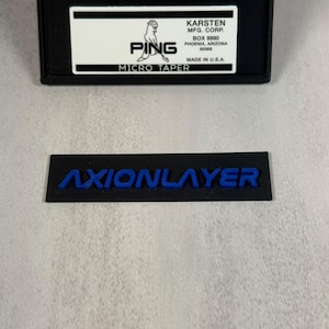 May include: Black rectangular box with a white label featuring the PING logo and text. Below is a black rectangle with the blue text "AXIONLAYER". The box also includes the text "MICRO TAPER" and "MADE IN U.S.A."