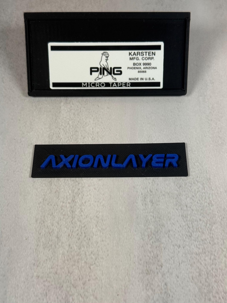 May include: Black rectangular box with a white label featuring the PING logo and text. Below is a black rectangle with the blue text "AXIONLAYER". The box also includes the text "MICRO TAPER" and "MADE IN U.S.A."