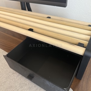 May include: A monitor stand featuring a black frame with a pull-out drawer and a top surface constructed from several parallel light-coloured wooden dowels. The word AXIONLAYER is printed in white on the front edge of the black drawer frame.
