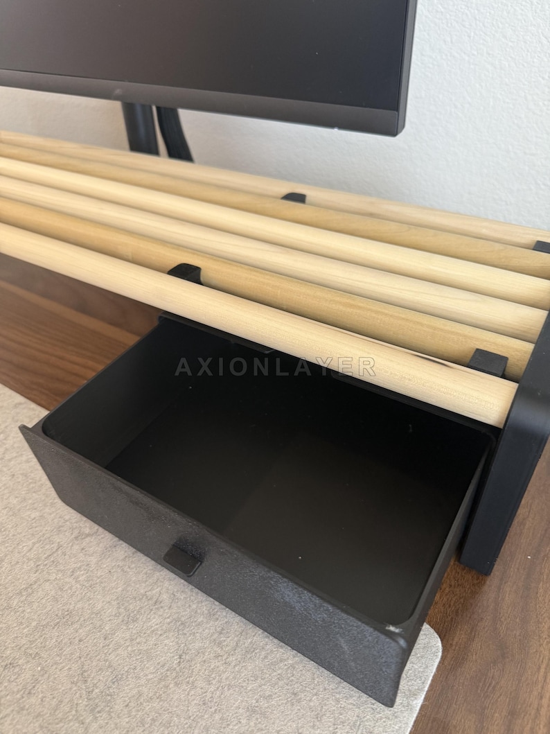 May include: A monitor stand featuring a black frame with a pull-out drawer and a top surface constructed from several parallel light-coloured wooden dowels. The word AXIONLAYER is printed in white on the front edge of the black drawer frame.