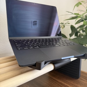 May include: A dark grey laptop with a black keyboard and trackpad is positioned on a black adjustable stand. The stand features a series of light-coloured wooden dowels across its surface, providing a stable base for the electronic device.