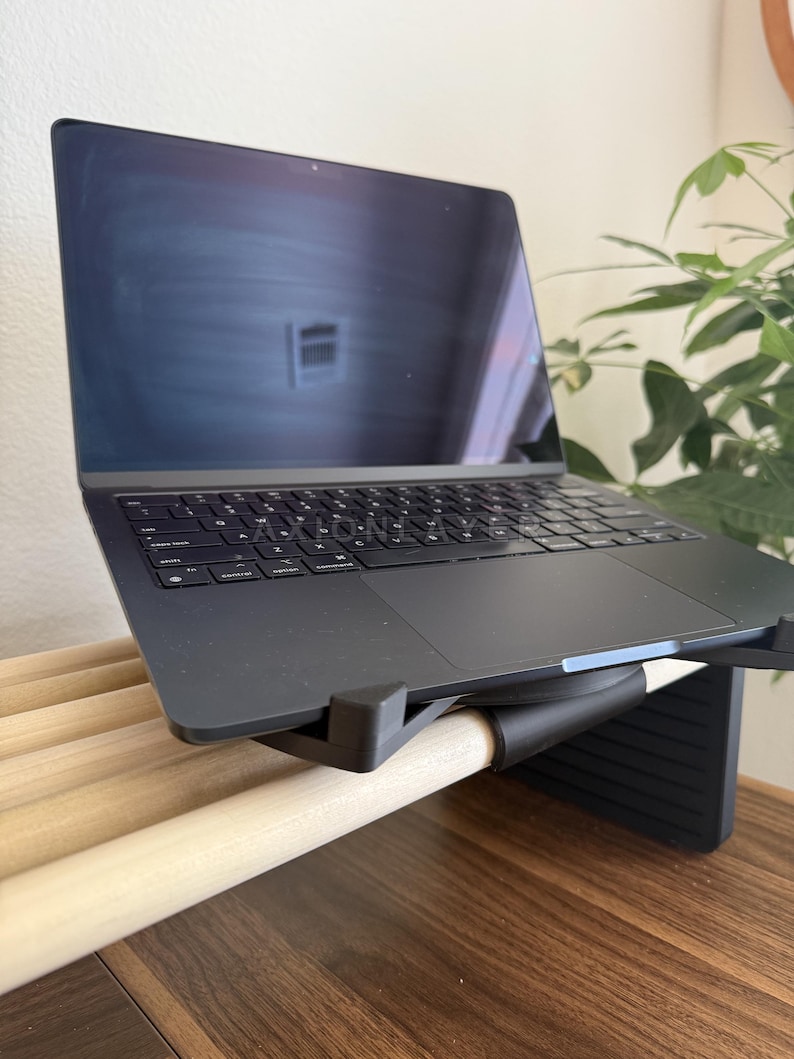 May include: A dark grey laptop with a black keyboard and trackpad is positioned on a black adjustable stand. The stand features a series of light-coloured wooden dowels across its surface, providing a stable base for the electronic device.