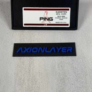 May include: Black rectangular label with the word "AXIONLAYER" in blue lettering. Above it is a black rectangular box with a white label that reads "PING" and "KARSTEN MFG. CORP." in red and black text. Made in the U.S.A.
