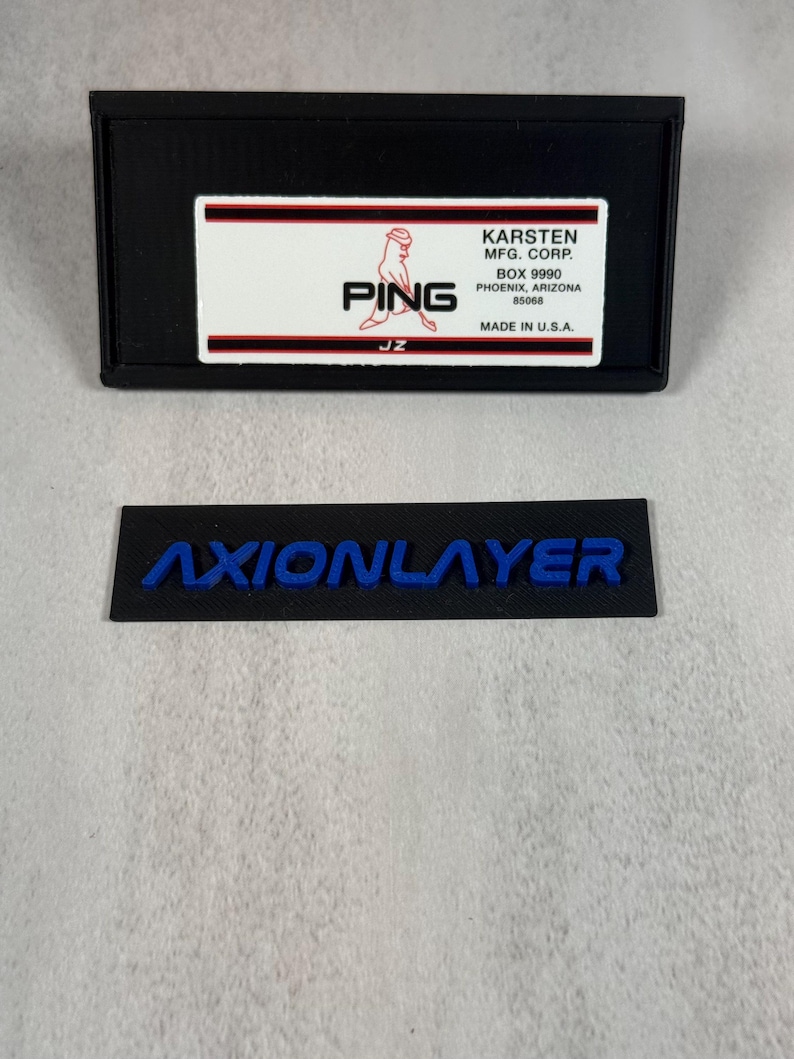 May include: Black rectangular label with the word "AXIONLAYER" in blue lettering. Above it is a black rectangular box with a white label that reads "PING" and "KARSTEN MFG. CORP." in red and black text. Made in the U.S.A.