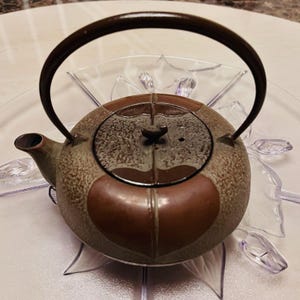 Puede incluir: Una tetera redonda de hierro fundido marrón y verde con un asa y tapa marrón oscuro. La tetera tiene un pico y está decorada con un diseño en forma de corazón. Se encuentra sobre un plato decorativo transparente.