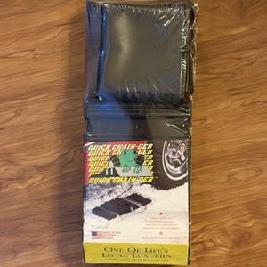 Puede incluir: Un producto automotriz negro y rectangular empaquetado en plástico transparente. El embalaje presenta el texto "QUICK CHAIN-GER" con un diseño de rana y una imagen de neumático. El producto está etiquetado como "ONE OF LIFE'S LITTLE LUXURIES."