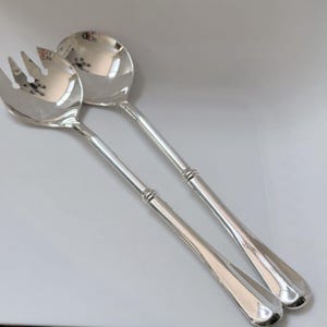 May include: A pair of polished silver-tone salad serving utensils consisting of a large serving spoon and a matching serving fork. Both pieces feature long, slender handles with a subtle triple-ring decorative detail near the neck of the bowl.
