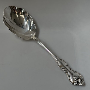 May include: A polished silver-toned serving spoon featuring a large, scalloped bowl and an ornate, embossed handle with intricate floral and scrollwork detailing. The metal surface is highly reflective, showing smooth curves and decorative relief.