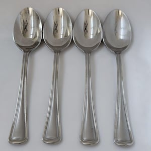 May include: Four silver spoons with oval bowls and long, tapered handles. The spoons are arranged in a row, reflecting light. The handles have a decorative, ridged design along the edges.