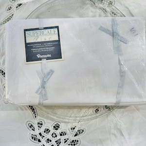 May include: A white Twin X-Long fitted sheet set, still in its original packaging. The label reads "Supercale Plus" and includes details about the thread count and material composition. The sheet set is tied with light blue ribbons.