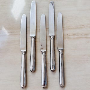 May include: A set of six silver-colored knives arranged on a light-colored surface. Each knife has a long, slender blade and a rounded handle. The knives are evenly spaced and appear to be made of metal, reflecting the light.