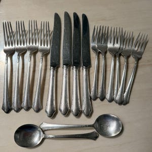 Könnte beinhalten: Eine Sammlung antiker Silberbestecke, darunter Gabeln, Messer und Löffel. Das silberfarbene Besteck weist verzierte Griffe auf. Die Gabeln und Messer sind in Reihen angeordnet, während die Löffel unten platziert sind. Ein klassisches Set für das Abendessen.