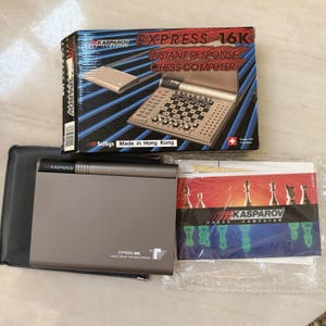 Puede incluir: Un juego de ajedrez electrónico Kasparov Express 16K que incluye un dispositivo portátil gris, una caja de cartón con imágenes y texto del producto, un estuche protector negro y un manual de instrucciones impreso con piezas de ajedrez.