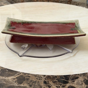 May include: A rectangular ceramic dish with a deep red centre and a wide, textured olive green border. The dish features a slightly curved, elevated design with a flared rim, resting on a clear, multi-sectioned plastic tray base.
