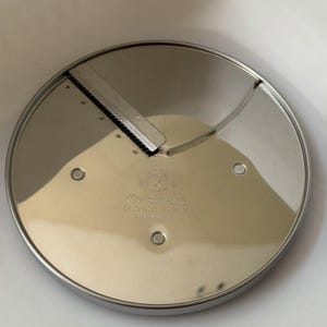 May include: A stainless steel Cuisinart DLC-032 slicing disc. The circular blade has a sharp, angled cutting edge and three small holes. The brand name and model number are printed on the surface.
