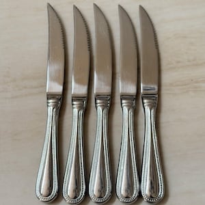 Pode incluir: Um conjunto de cinco facas de carne em aço inoxidável prateado dispostas verticalmente. Cada faca apresenta uma lâmina serrilhada e um cabo com uma borda decorativa de contas ao longo das laterais, terminando numa base arredondada.