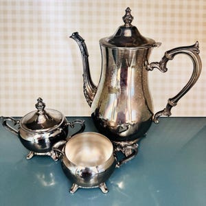 May include: A silver tea set with a coffee pot, sugar bowl, and creamer. The coffee pot has a curved handle and a decorative lid. The sugar bowl has a lid and two handles. The creamer has two handles. The set is on a blue surface.