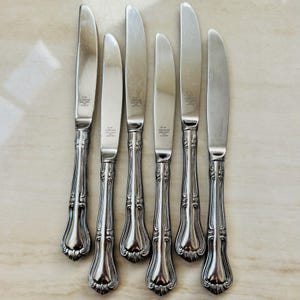May include: Set of seven silver-colored knives with ornate, detailed handles. The blades are smooth and shiny, and the handles feature a decorative, curved design. The knives are arranged on a light-colored surface, showcasing their elegant form.
