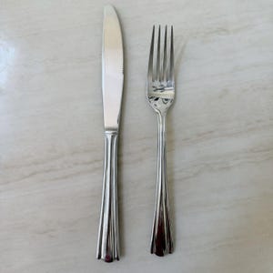May include: A stainless steel dinner knife and a matching four-tined dinner fork. Both pieces of cutlery feature a polished, reflective finish and a handle design characterized by three vertical, parallel grooves running down the center.
