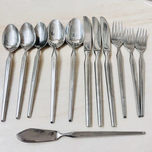 Könnte beinhalten: Eine Sammlung von Edelstahlbesteck, darunter Löffel, Messer, Gabeln und ein Buttermesser. Das Besteck hat ein schlichtes, modernes Design mit langen, schlanken Griffen. Das Besteck ist auf einer hellen Oberfläche angeordnet.