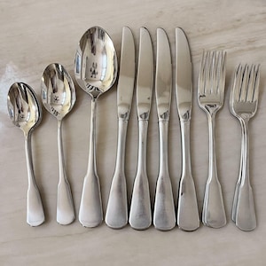 May include: An assortment of polished silverware, including spoons, knives, and forks, arranged on a light-colored surface. The silverware has a reflective, metallic sheen, suggesting stainless steel or silver. The arrangement showcases the various utensils used for dining.