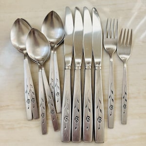 Op de afbeelding: Een verzameling roestvrijstalen bestek, waaronder lepels, messen en vorken. Elk stuk heeft een decoratief bloemen- en bladerontwerp dat in de handvatten is geëtst. Het bestek is gerangschikt op een lichtgekleurd oppervlak.