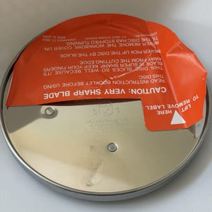 May include: A stainless steel Cuisinart DLC-7 slicing disc with an orange warning label. The label reads "CAUTION: VERY SHARP BLADE" and provides safety instructions. The disc is made in Japan.
